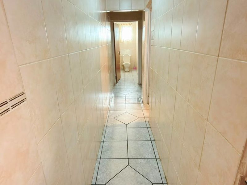 A narrow hallway featuring full-height ceramic wall tiling and patterned ceramic floor tiles, leading towards a bathroom.