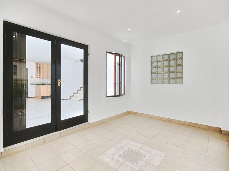 A bright, empty enclosed patio or sunroom featuring tiled flooring, a decorative glass block window, and double doors leading to the main living area.