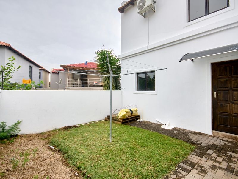 A rear exterior view of the property featuring a small lawn area, paved walkway, boundary wall, and an external air conditioning unit.