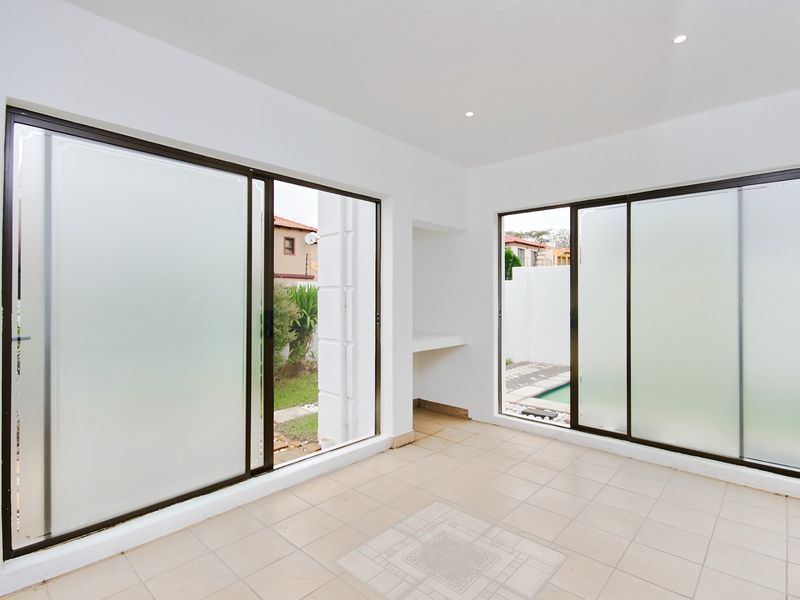 A bright, empty enclosed patio featuring tiled flooring, recessed lighting, and large sliding glass doors providing access to the garden and pool area.