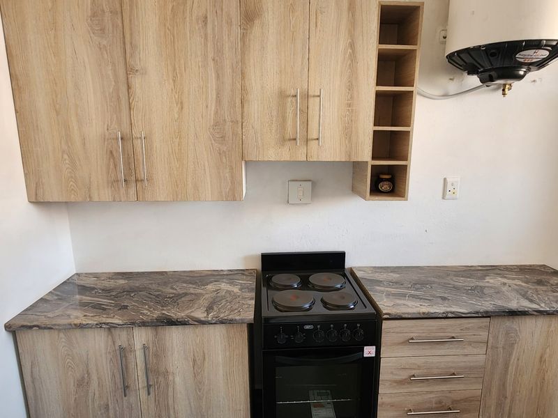 A compact kitchen featuring light wood-grain cabinetry, stone-look countertops, and a freestanding electric stove, with an exposed wall-mounted water heater.