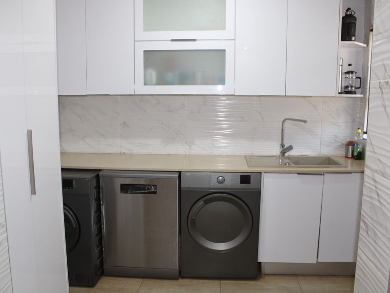 A modern, well-maintained laundry area featuring white cabinetry, stone countertops, marble-look wall tiles, and a stainless steel sink.