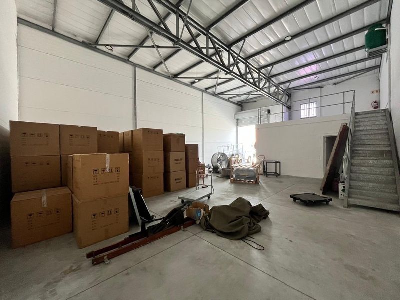 Spacious industrial warehouse unit featuring high ceilings with exposed steel trusses, concrete flooring, a mezzanine level, and a roller shutter door for easy access.
