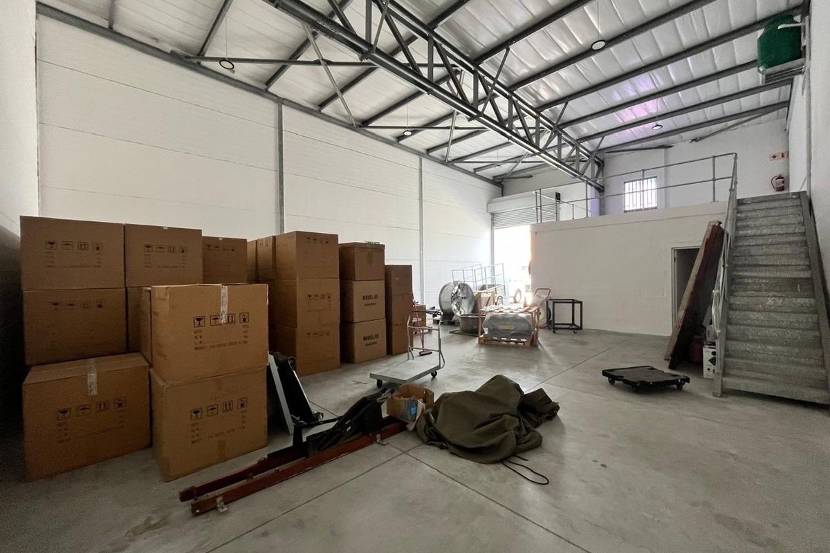Spacious industrial warehouse unit featuring high ceilings with exposed steel trusses, concrete flooring, a mezzanine level, and a roller shutter door for easy access.