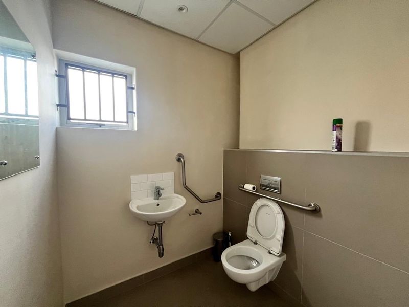 A functional, accessible bathroom featuring a wall-mounted toilet, basin, and safety grab rails, finished with neutral tiling and a drop ceiling.