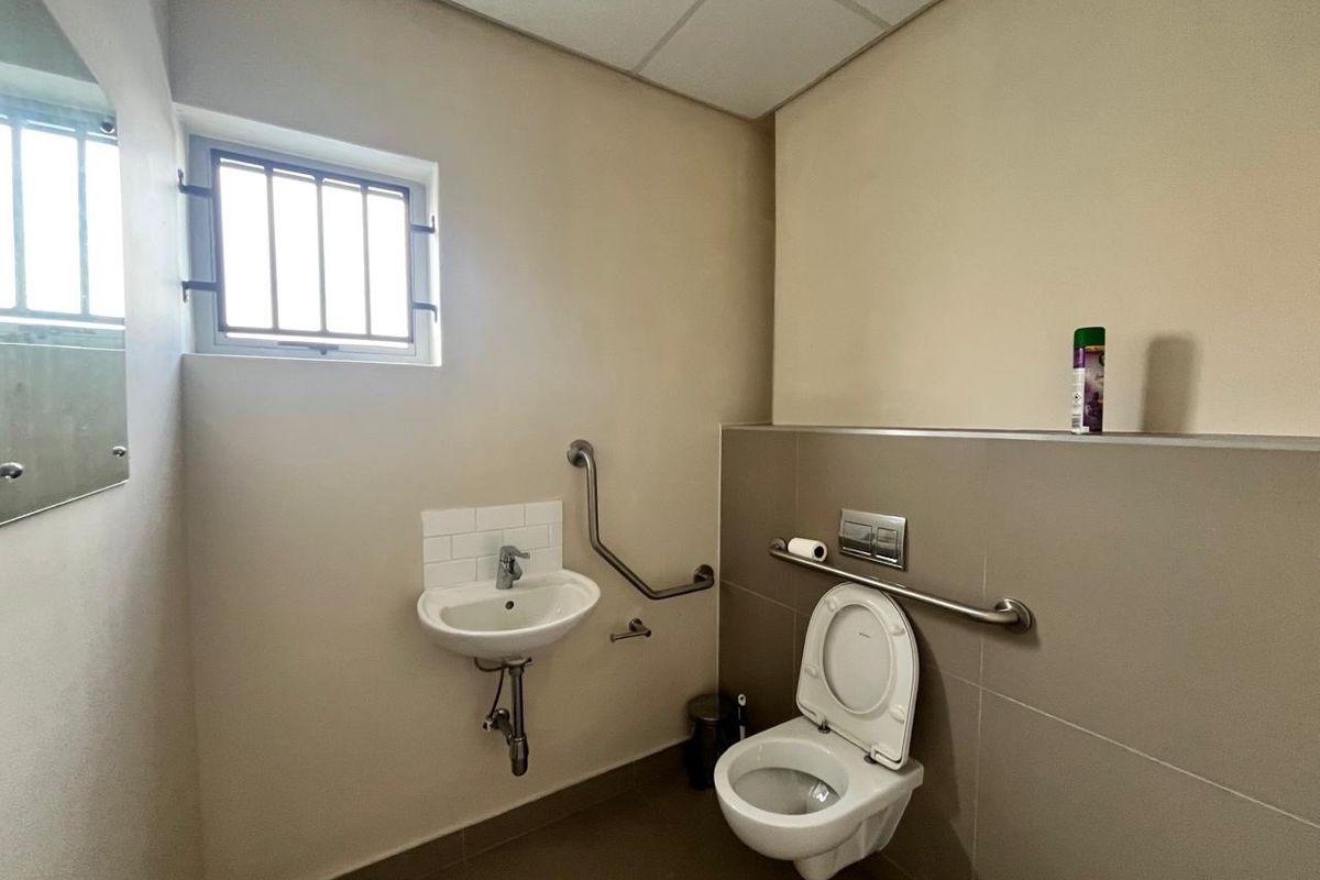 A functional, accessible bathroom featuring a wall-mounted toilet, basin, and safety grab rails, finished with neutral tiling and a drop ceiling.