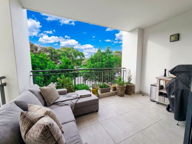 A spacious covered balcony featuring neutral floor tiles and a metal balustrade, offering an open outlook with pleasant greenery.