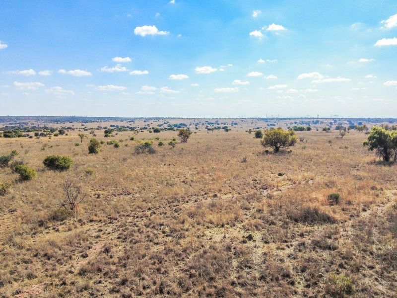 An aerial view of a large, undeveloped residential plot featuring natural grassland and scattered native trees under a clear blue sky.