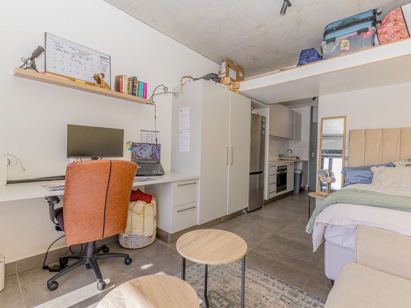 A modern open-plan studio apartment featuring an exposed concrete ceiling, built-in desk, integrated kitchen cabinetry, and a functional loft storage space.