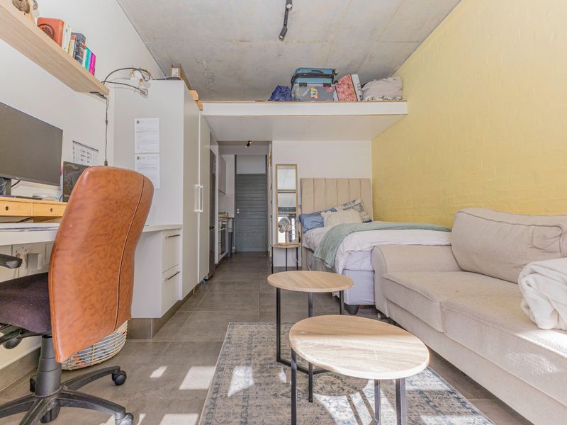 Compact open-plan studio apartment featuring a mezzanine level, exposed concrete ceiling, tiled flooring, and a functional built-in desk area.