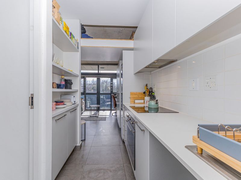 Modern galley-style kitchen featuring sleek white cabinetry, integrated appliances, stone countertops, and a tiled backsplash, leading towards an open-plan living area.