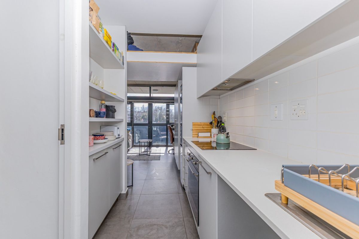Modern galley-style kitchen featuring sleek white cabinetry, integrated appliances, stone countertops, and a tiled backsplash, leading towards an open-plan living area.