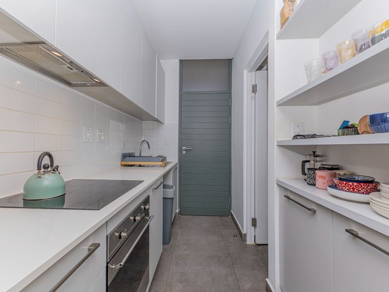 A compact, modern scullery featuring white cabinetry, an electric hob, built-in oven, extractor fan, and practical open shelving for additional storage.
