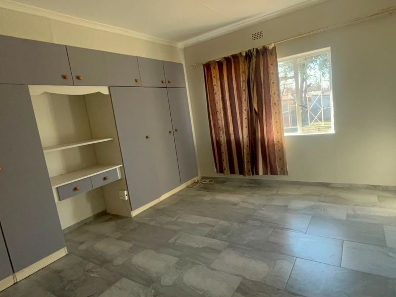 A spacious bedroom featuring built-in cupboards with a vanity area, tiled flooring, and a window fitted with security bars.