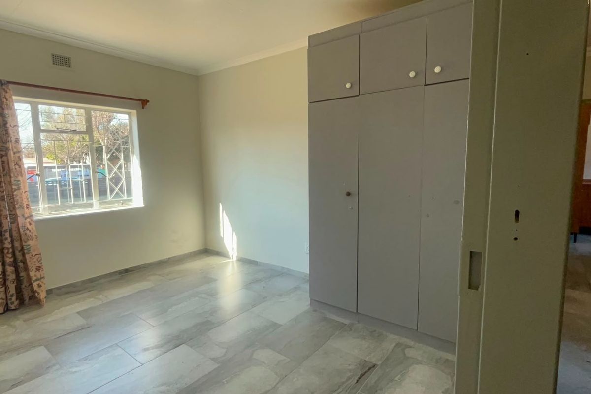 A bright, empty bedroom featuring neutral tiled flooring, built-in cupboards, and a window with security bars.