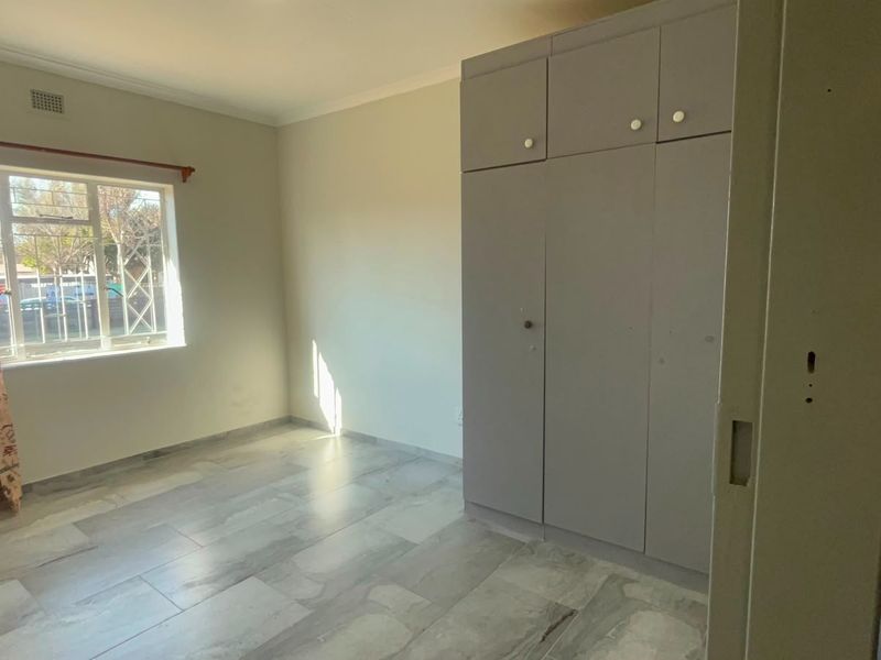 A bright, empty bedroom featuring neutral tiled flooring, built-in cupboards, and a window with security bars.
