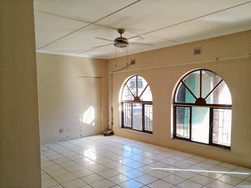 An empty living room featuring arched windows with security bars, tiled flooring, and a ceiling fan, requiring cosmetic updates to improve its appeal.
