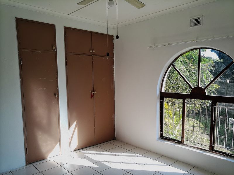 An empty bedroom featuring tiled flooring, built-in cupboards, a ceiling fan, and a distinctive arched window with security bars.