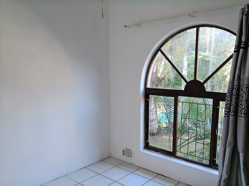 An empty bedroom featuring a distinctive arched window with security bars and tiled flooring, offering a view of the garden.