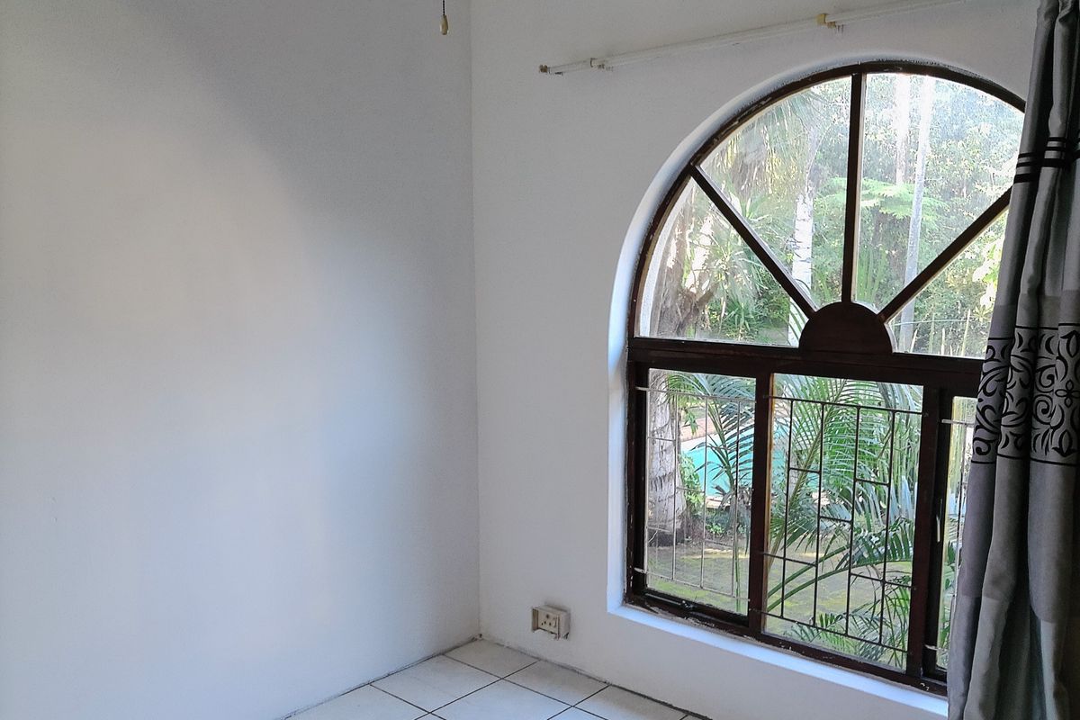 An empty bedroom featuring a distinctive arched window with security bars and tiled flooring, offering a view of the garden.