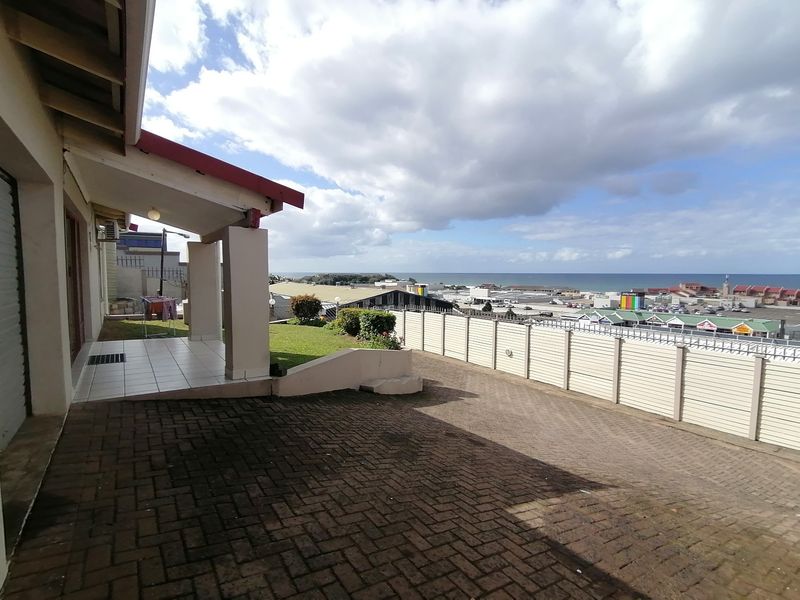 Rear exterior view featuring a paved driveway, covered patio area, and a lawn with a boundary wall overlooking the coastal town and ocean.