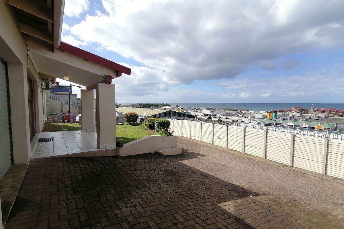 Rear exterior view featuring a paved driveway, covered patio area, and a lawn with a boundary wall overlooking the coastal town and ocean.