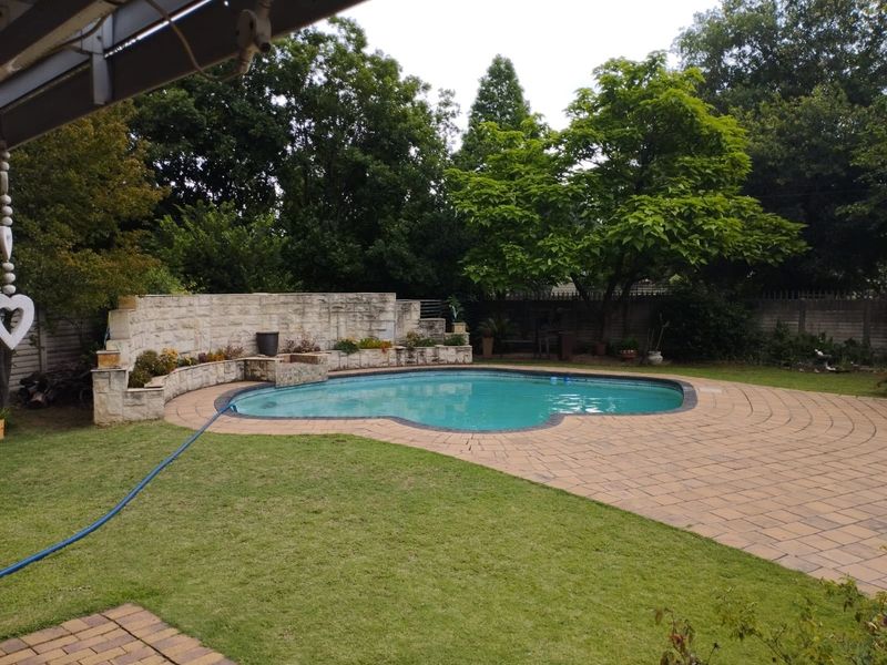 A well-maintained outdoor pool area featuring a curved stone retaining wall, paved pool deck, and established garden surroundings.