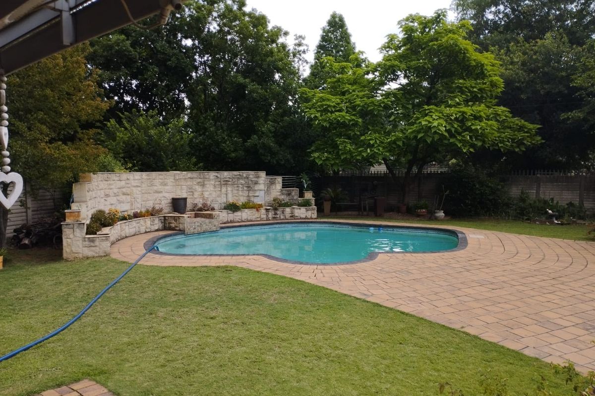 A well-maintained outdoor pool area featuring a curved stone retaining wall, paved pool deck, and established garden surroundings.
