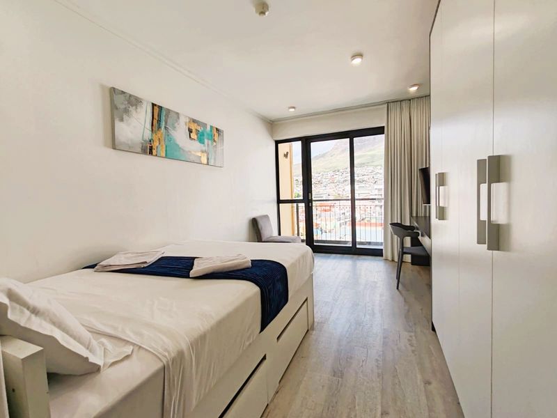 A bright bedroom featuring built-in wardrobes, laminate flooring, and a sliding glass door leading to a balcony with scenic views.