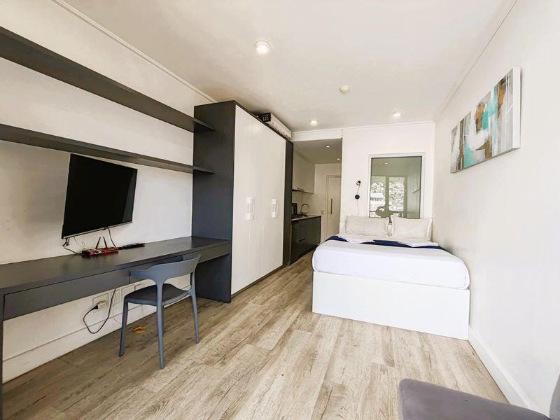 A modern open-plan studio apartment featuring a built-in desk, ample wardrobe storage, and a compact kitchenette, finished with light wood-look flooring.
