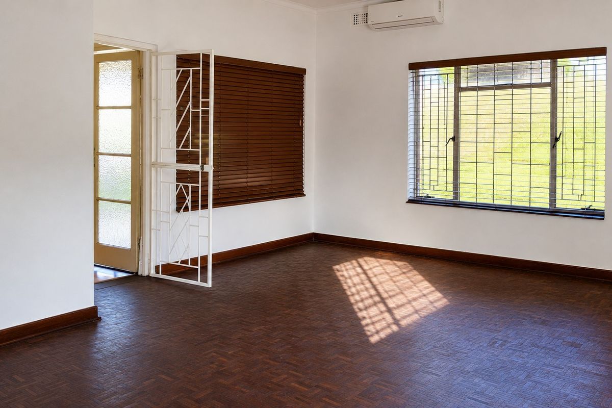 A bright, empty living room featuring classic parquet flooring, an air conditioning unit, and secure window treatments.