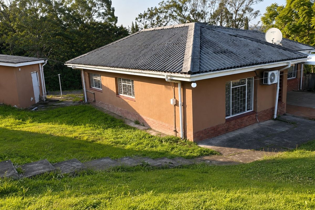 Rear view of a detached house featuring a corrugated roof, brick and plaster facade, security bars on windows, and a sloped garden with concrete stairs.