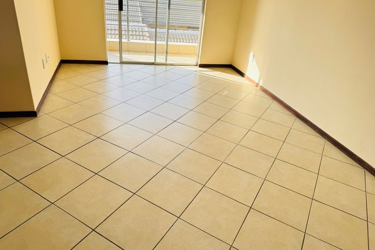 An empty, sunlit living area featuring neutral ceramic floor tiles and a sliding glass door providing access to an outdoor balcony.