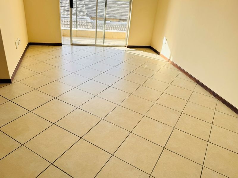An empty, sunlit living area featuring neutral ceramic floor tiles and a sliding glass door providing access to an outdoor balcony.