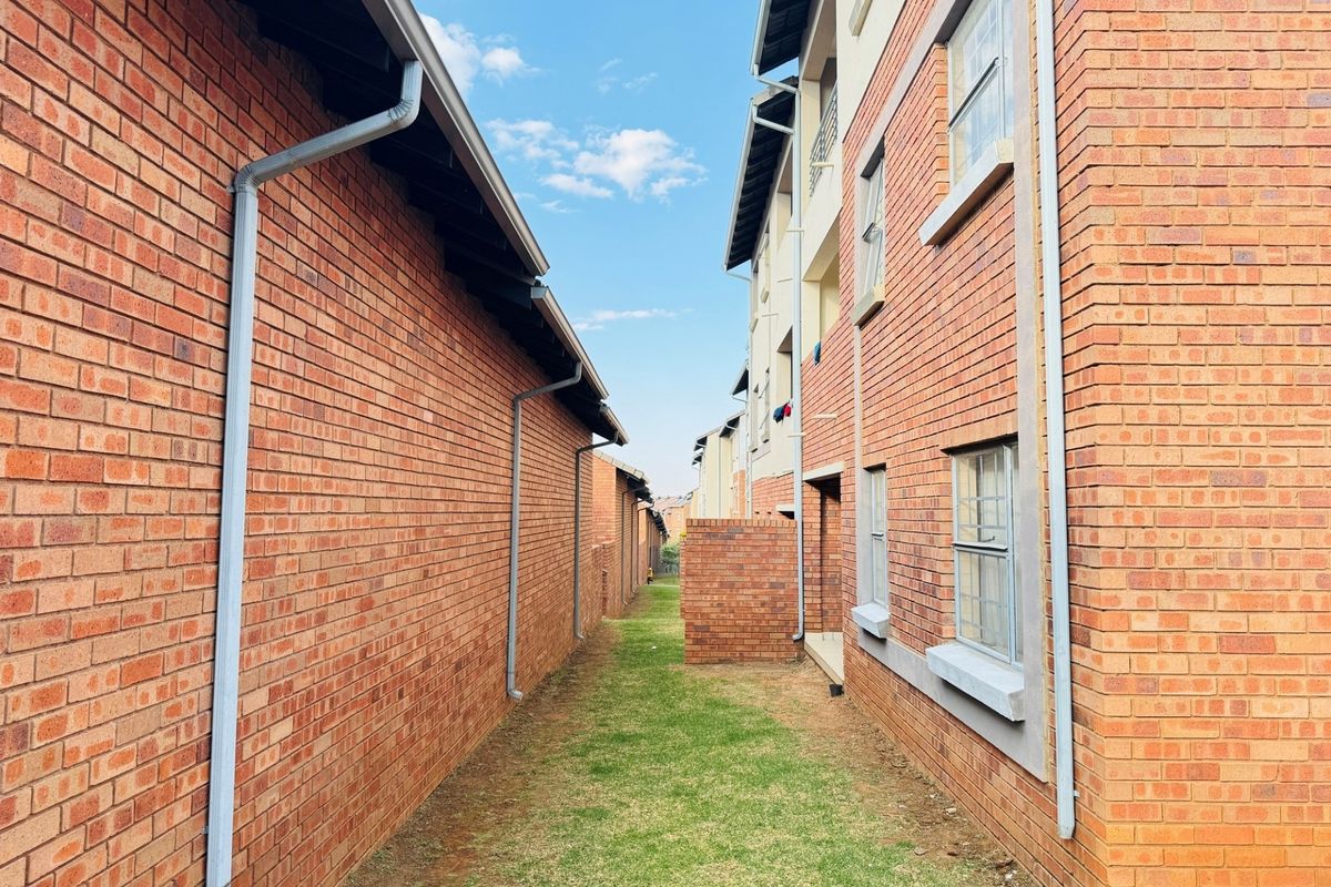 A view of the side passage between apartment blocks, featuring face brick exterior walls, installed rainwater downpipes, and a small grassed area.