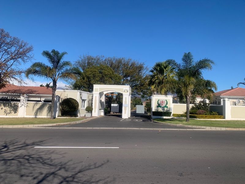 Street view of the gated entrance to the residential estate, featuring a paved driveway, perimeter walling, and established palm trees.