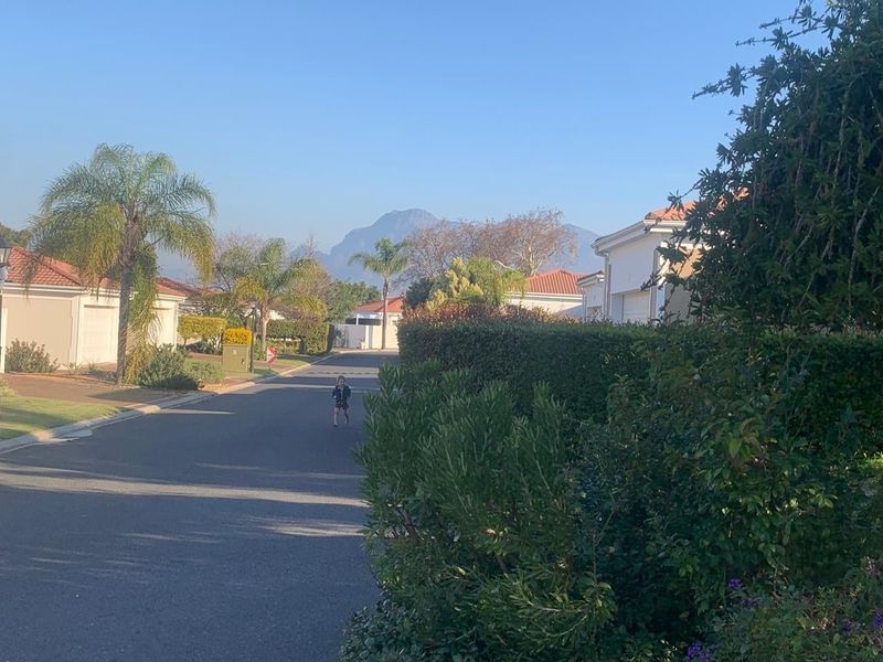 A view of a residential street within a well-maintained estate, featuring landscaped hedges, tiled roof homes, and mountain views in the background.