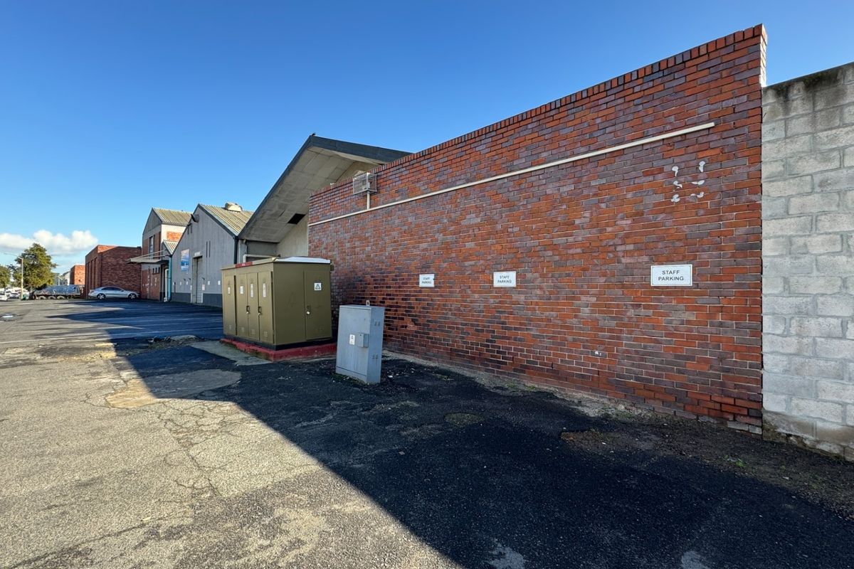 Rear exterior view of an industrial property featuring a brick facade, asphalt parking area, and an external electrical substation.