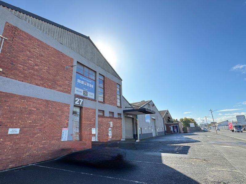 Front elevation of an industrial warehouse property featuring brick construction, a roller shutter door, and an expansive asphalt parking area.