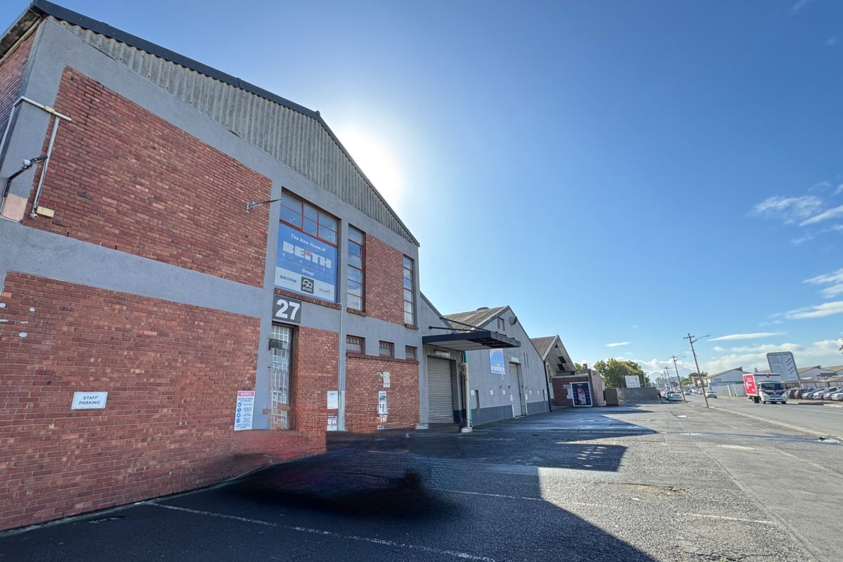 Front elevation of an industrial warehouse property featuring brick construction, a roller shutter door, and an expansive asphalt parking area.