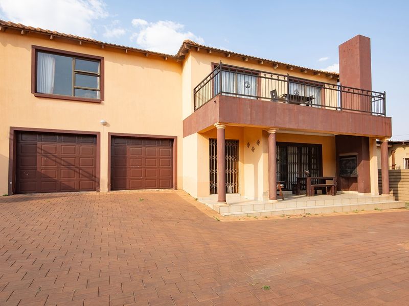Front elevation of a double-story townhouse featuring a double garage, paved driveway, and an upper-level balcony with a built-in braai area.