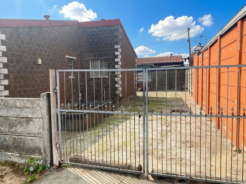 Rear exterior view showing a paved driveway, metal security gate, and surrounding brick and precast concrete boundary walls.