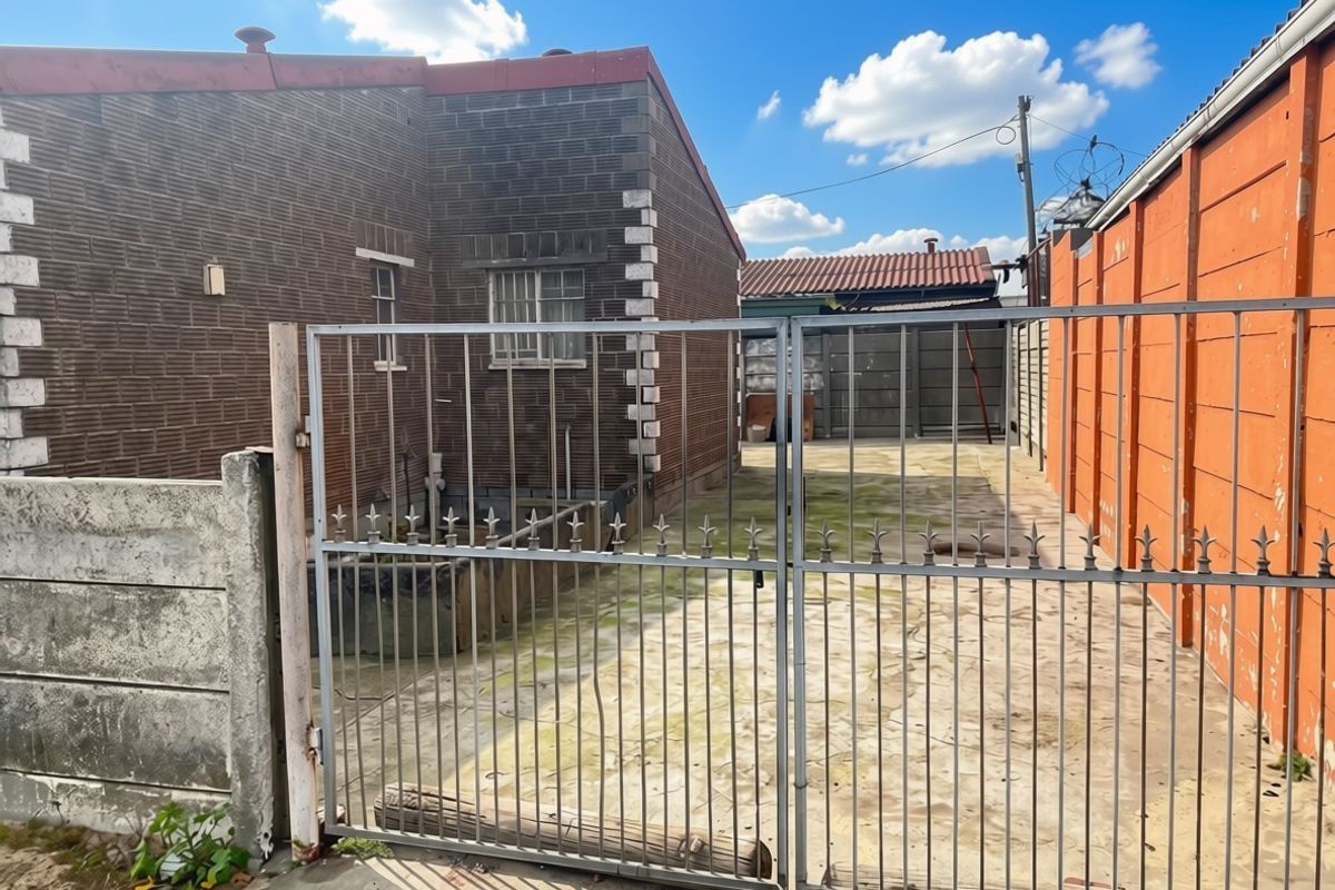 Rear exterior view showing a paved driveway, metal security gate, and surrounding brick and precast concrete boundary walls.