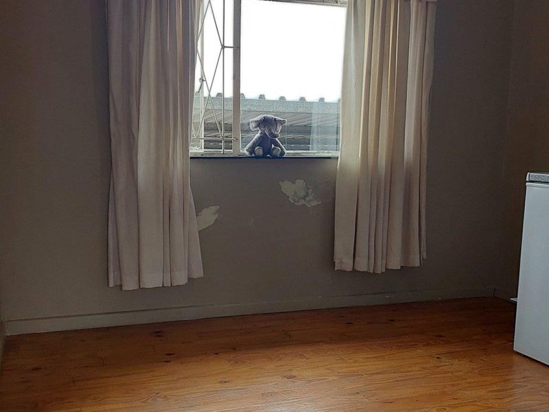 A sparsely furnished bedroom featuring timber flooring and a window with security bars, showing signs of peeling paint on the wall.