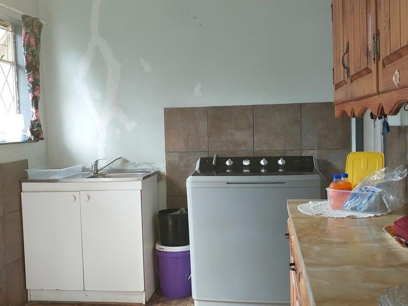 A functional laundry area featuring a stainless steel sink, tiled splashback, and wooden cabinetry, currently showing signs of wear and wall damage.