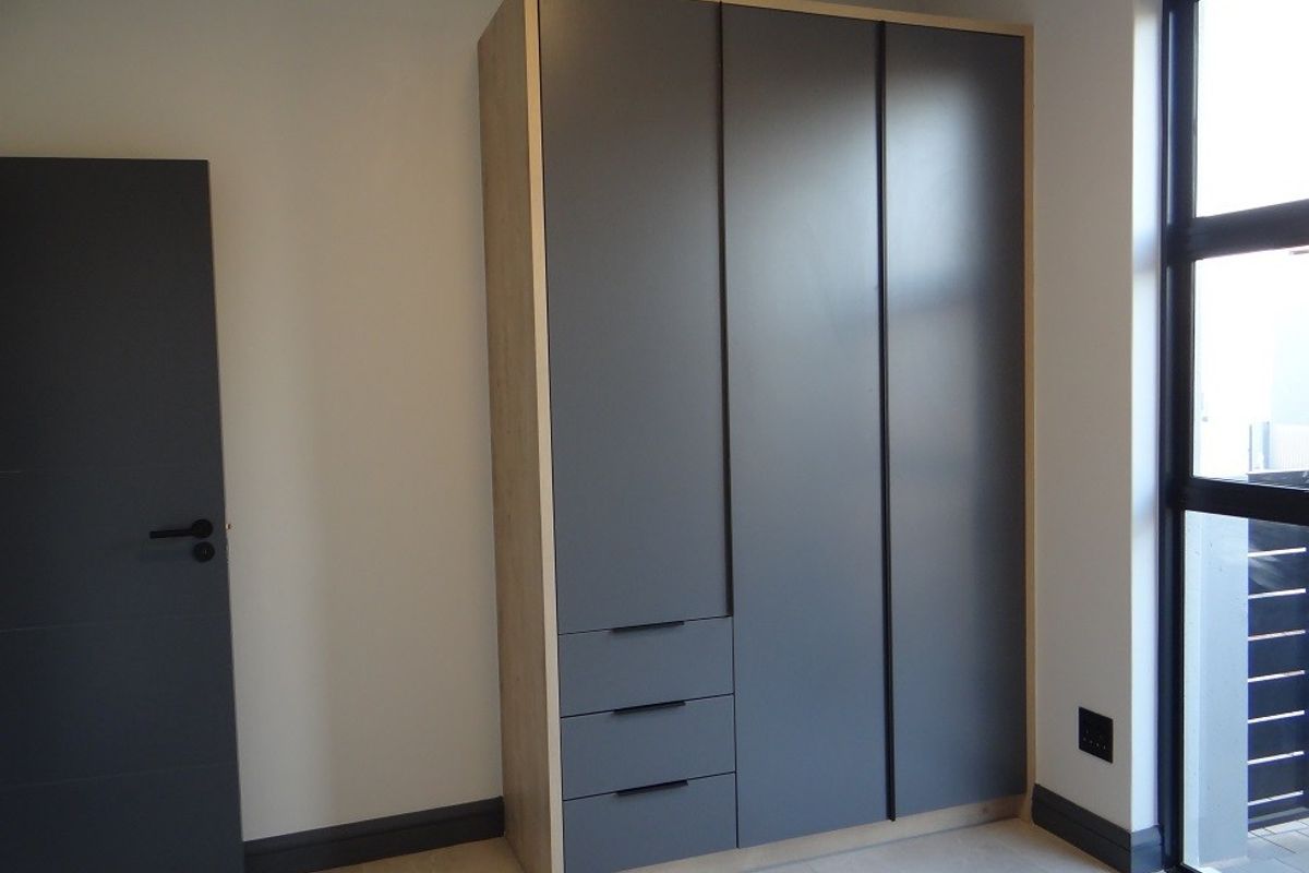 A modern bedroom featuring a sleek, built-in wardrobe with grey cabinetry, light wood trim, tiled flooring, and a large black-framed window.