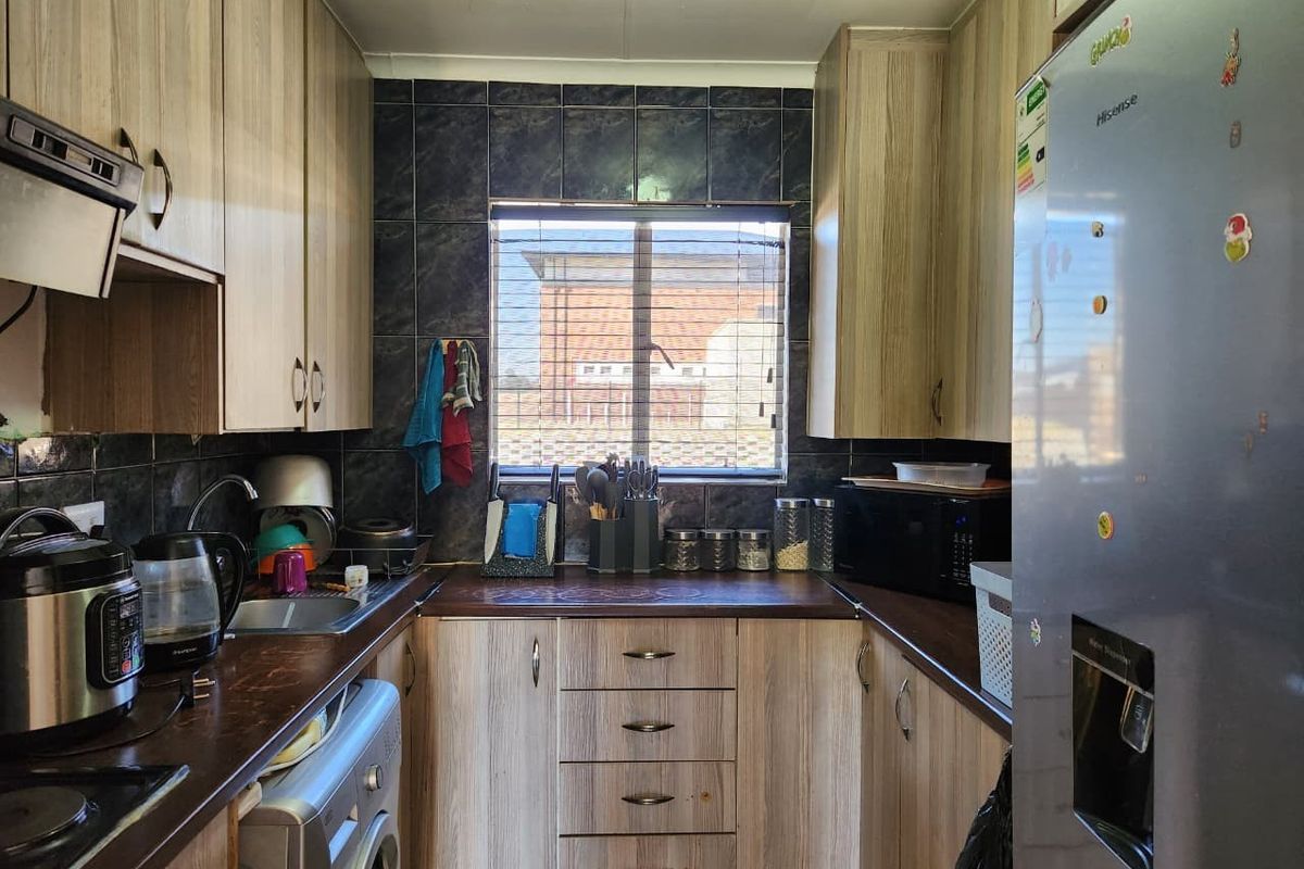 Compact galley-style kitchen featuring wood-look laminate cabinetry, dark countertops, tiled backsplash, and integrated plumbing for laundry appliances.