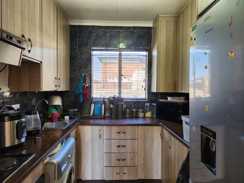 Compact galley-style kitchen featuring wood-look laminate cabinetry, dark countertops, tiled backsplash, and integrated plumbing for laundry appliances.