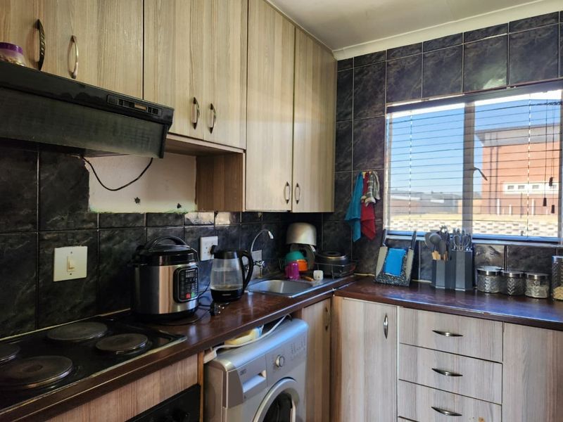 Compact kitchen featuring wood-look cabinetry, dark tiled backsplash, electric hob, and integrated laundry plumbing, requiring minor cosmetic repairs to the wall and tiling.