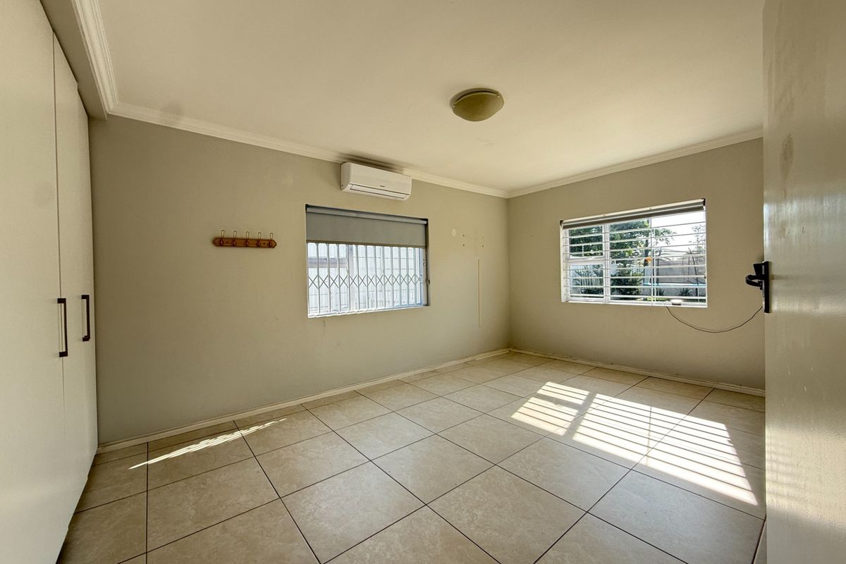 A bright, empty bedroom featuring tiled flooring, built-in cupboards, an air conditioning unit, and security bars on the windows.